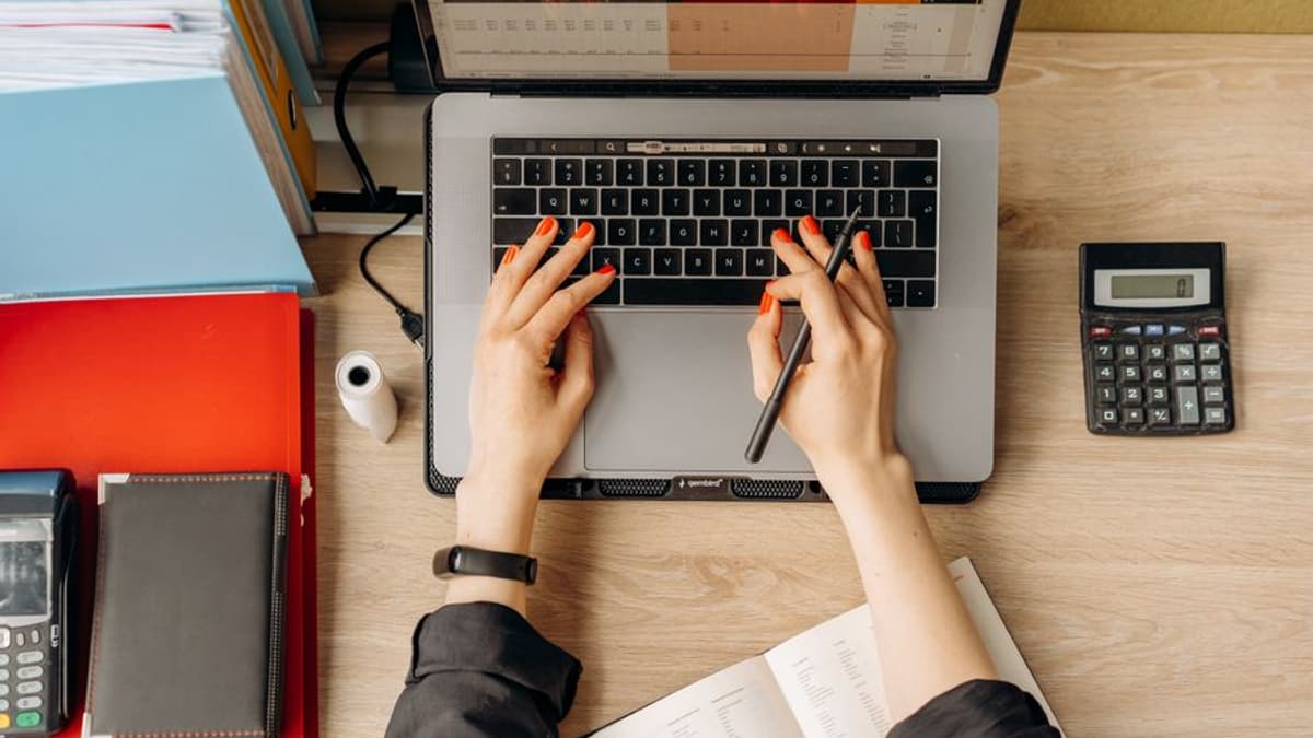 Mãos digitando em um notebook com uma planilha na tela e uma calculadora ao lado